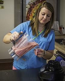 A woman in a blue shirt pouring a pink smoothie from a blender into a glass. Links to the Nutrition Corner page.  