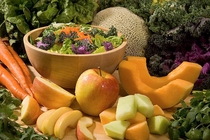 A bowl of salad greens surrounded by slices apples, cantaloupe slices, whole cantaloupes, whole carrots and kale