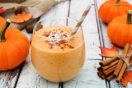 A pumpkin smoothie in a glass with a straw sitting on wood and surrounded by pumpkins and cinnamon sticks. 