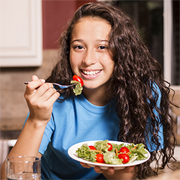 A young girl wearing a blue shirt and eating a plate of salad. 