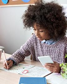 An African American girl sitting at a desk holding a mobile phone while writing notes 