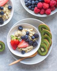 Two bowls of oatmeal topped with blueberries and strawberries surrounded by sliced kiwi fruit. 