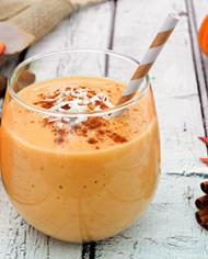A pumpkin smoothie in a glass with a straw sitting on wood and surrounded by pumpkins and cinnamon sticks. 