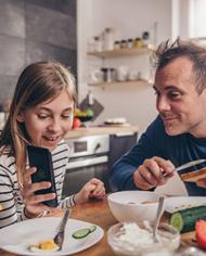 Father and daughter sitting at a kitchen table looking at a smart phone while eating