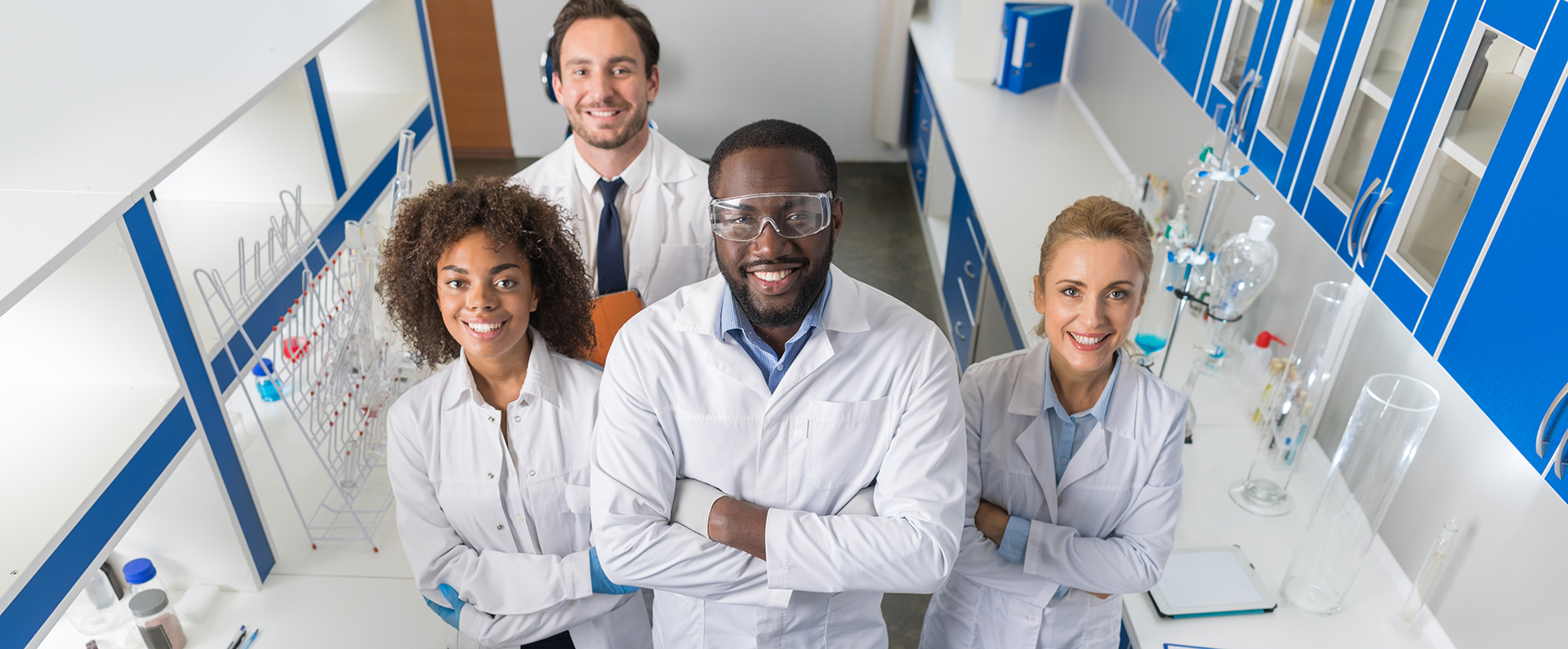 Two women and two men wearing white lab coats and standing with their arms crossed in a room of beakers and test tubes