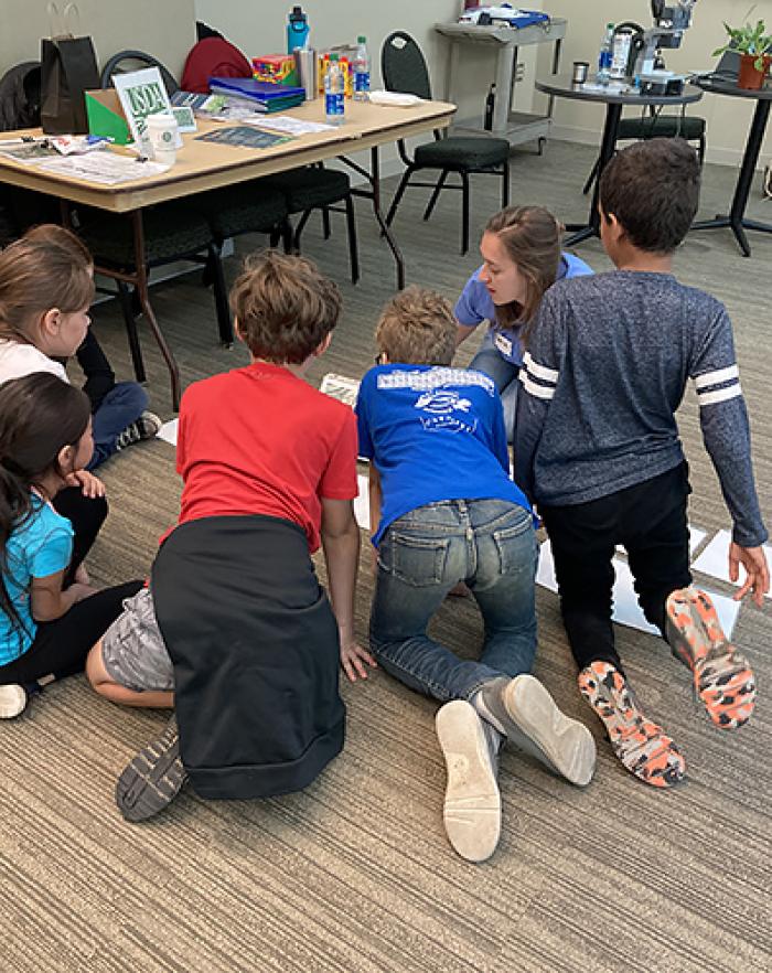 ARS researcher Hanna Oleszak sitting on the floor talking with six students grouped around her. 
